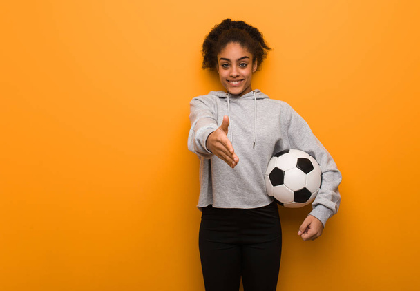 Молодая фитнес черная женщина протягивает руку, чтобы поприветствовать кого-то. Холдинг футбольный мяч
. - Фото, изображение