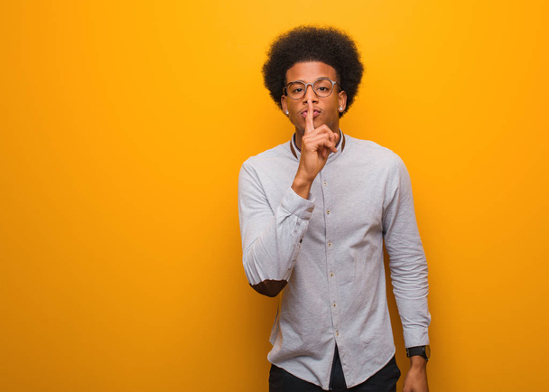 Fiatal afro-amerikai férfi egy narancssárga falon, aki titkot tart, vagy csendet kér. - Fotó, kép