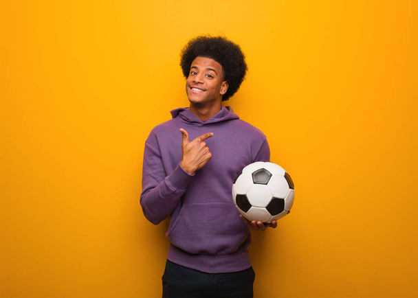 Молодой африканский американский спортсмен держит футбольный мяч, приглашая прийти
 - Фото, изображение