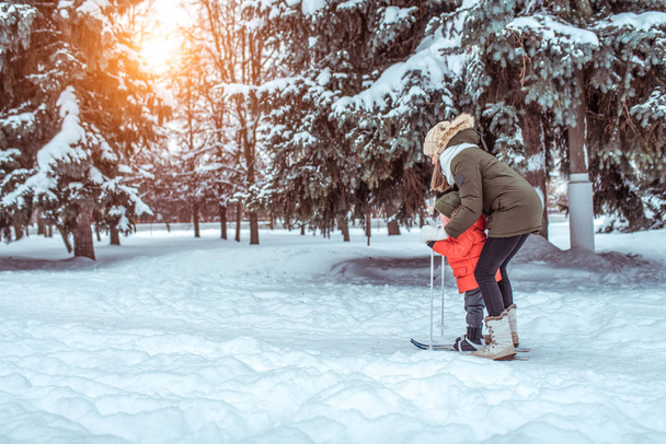A young mother supports a young son, a boy of 3 years. In winter, outside in park, background is snow drifts of Christmas tree. Free space. Childrens skis, skiing lessons, first steps, child care. - Zdjęcie, obraz