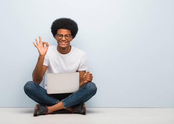 Joven hombre negro sentado en el suelo con un ordenador portátil alegre y seguro haciendo buen gesto
 - Foto, Imagen