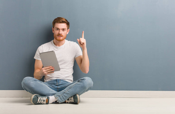 Young redhead student man sitting on the floor showing number one. He is holding a tablet. - Foto, Bild