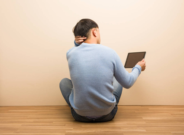 Young chinese man sitting using his tablet from behind thinking about something - Photo, Image