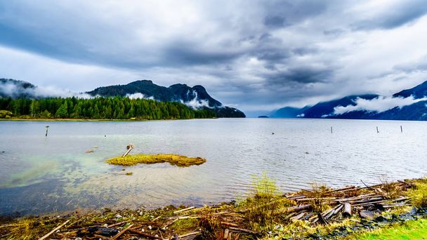 カナダ、ブリティッシュコロンビア州のフレイザーバレーの海岸山脈の山々にぶら下がる雨雲と暗い曇りの空の下でピット湖の海岸に流木 - 写真・画像