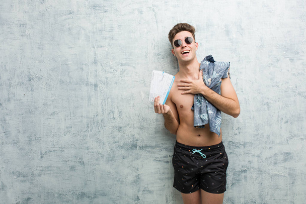 Een jongeman in een zwempak met vliegtickets lacht luid, hand in hand.. - Foto, afbeelding