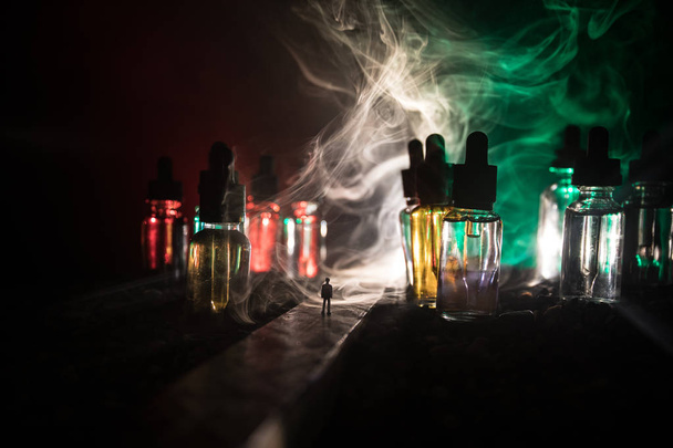 vape Konzept. Silhouette eines Mannes, der in einer nebligen Nacht mitten auf der Straße steht, mit riesigen Glasflaschen gefüllt mit Flüssigkeit für elektronische Zigaretten. bunte Nebelwolken mit Licht auf dem Hintergrund. - Foto, Bild