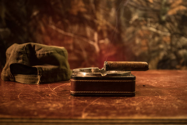 Primer plano de un cigarro cubano y cenicero sobre la mesa de madera. Mesa de comandante dictador comunista en cuarto oscuro. Concepto de mesa de trabajo del general del ejército
. - Foto, imagen