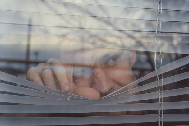 Чоловік дивиться через віконні жалюзі. Портрет молодого вдумливого чоловіка з коричневими очима, що спостерігають через вікно ревнощів
 - Фото, зображення