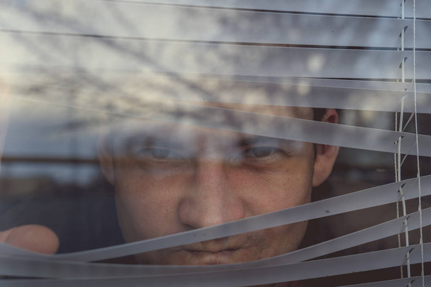 Man watching through window blinds. Portrait of young thoughtful male with brown eyes observing through window jalousie - Photo, image