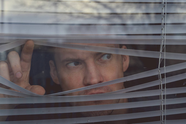 Man watching through window blinds. Portrait of young thoughtful male with brown eyes observing through window jalousie - Foto, Bild