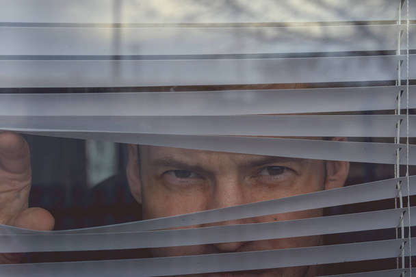 Mann schaut durch Jalousien. Porträt eines jungen, nachdenklichen Männchens mit braunen Augen, das durch Fenster Jalousie beobachtet - Foto, Bild