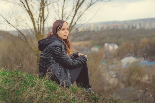 Young woman sitting on green hill. Side view of young dreamy female sitting on altitude and looking at suburb in spring day - Fotoğraf, Görsel