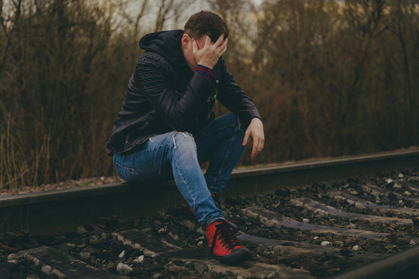 Desperate man sitting on railroad. Side view of hopeless male in casual outfit touching head while sitting on rails in autumn countryside - Фото, изображение