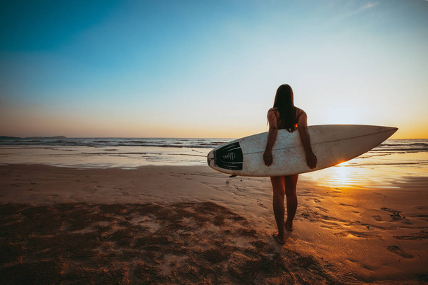 Surfer Kobieta w bikini przejść do surfingu. Sylwetka pięknej seksownej kobiety z deski surowej na plaży o zachodzie słońca. rocznika kolor ton - Zdjęcie, obraz
