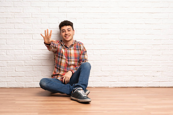 Νέος άνθρωπος κάθεται στο πάτωμα μετρώντας πέντε με τα δάχτυλα - Φωτογραφία, εικόνα