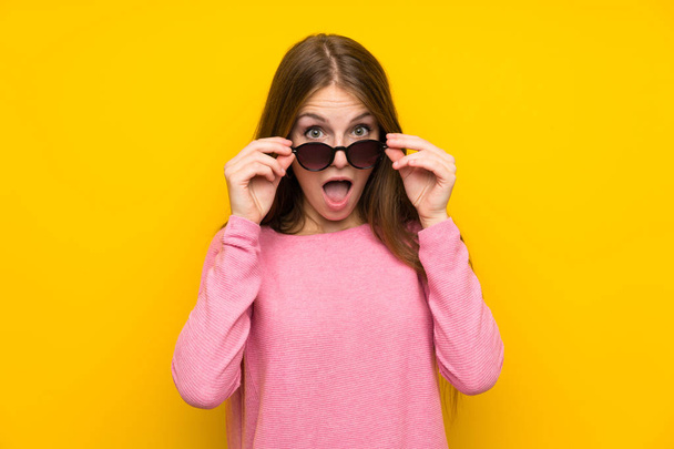 Young woman with long hair over isolated yellow wall with glasses and surprised - Foto, Imagen