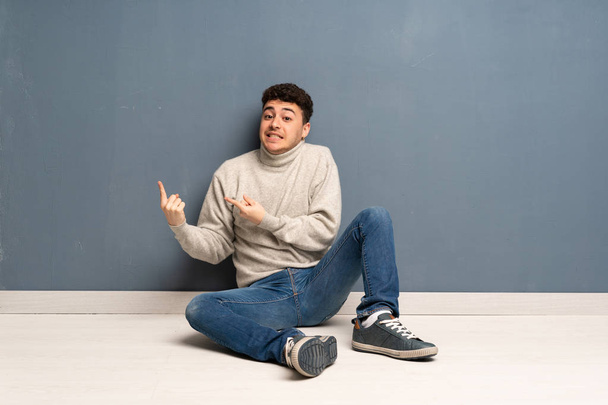Young man sitting on the floor frightened and pointing to the side - Fotoğraf, Görsel