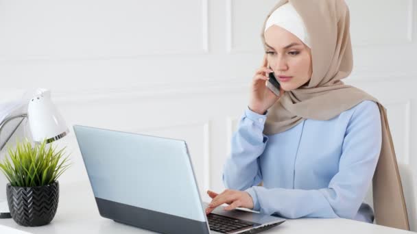 Muzułmańska kobieta w hidżab mówi telefon komórkowy i wpisując na laptopie w biurze pracy. - Materiał filmowy, wideo