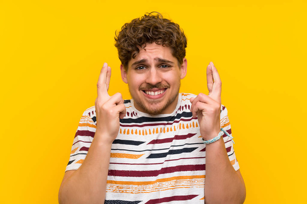 Blonde man over yellow wall with fingers crossing - Photo, Image