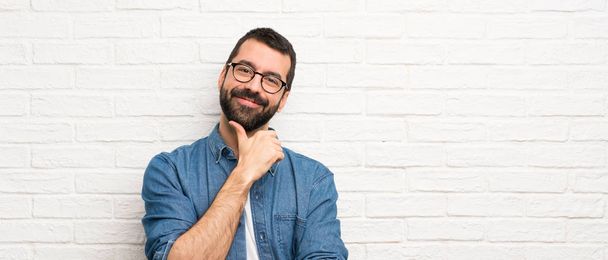 Pohledný muž s plnovousem přes bílou cihlovou zeď s brýlemi a s úsměvem - Fotografie, Obrázek