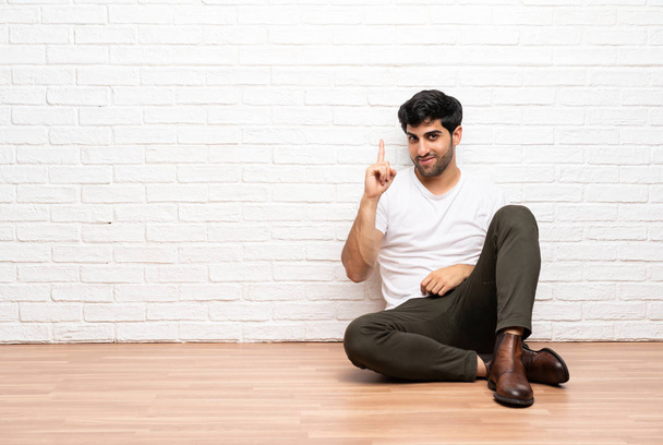 Young man sitting on the floor showing and lifting a finger in sign of the best - Foto, afbeelding