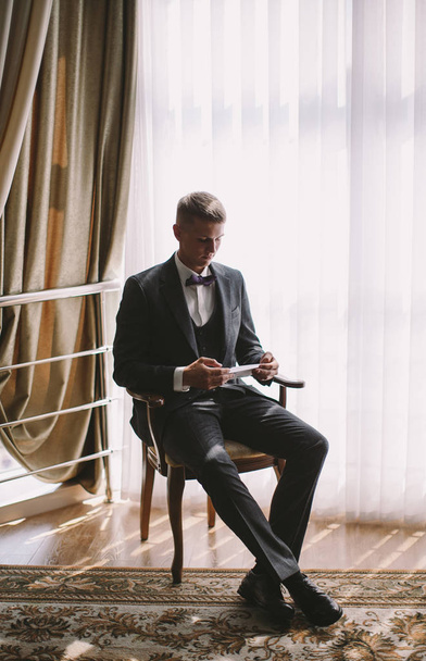 morning stylish groom - a young guy blond in a gray jacket reading a letter from the bride in a vintage room - Photo, image