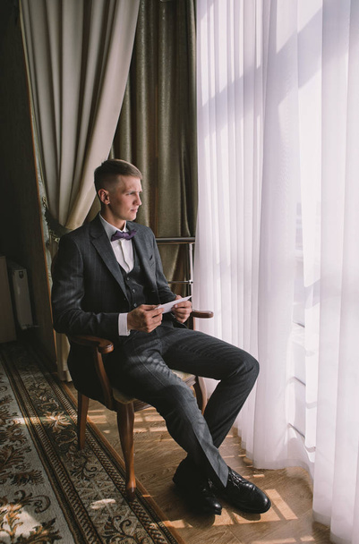朝のスタイリッシュな新郎 - ヴィンテージルームで花嫁からの手紙を読んで灰色のジャケットでブロンドの若い男 - 写真・画像