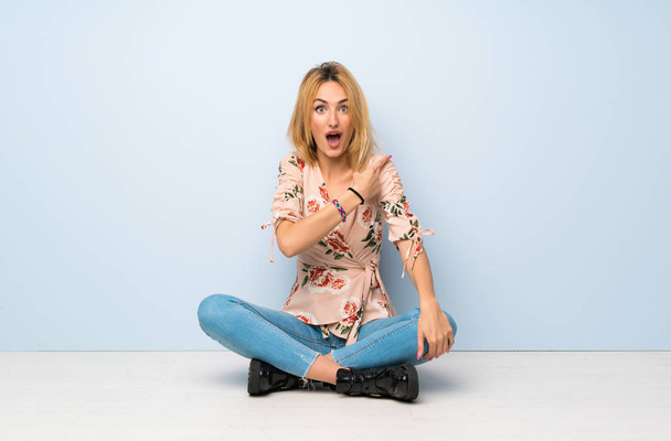 Mladá blondýnka sedí na podlaze překvapená a ukazuje stranou - Fotografie, Obrázek