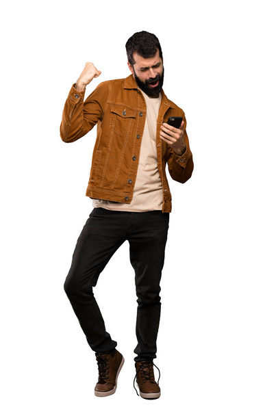 Handsome man with beard with phone in victory position over isolated white background - Valokuva, kuva