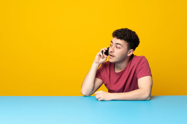 Joven con pared colorida y mesa manteniendo una conversación con el teléfono móvil
 - Foto, Imagen