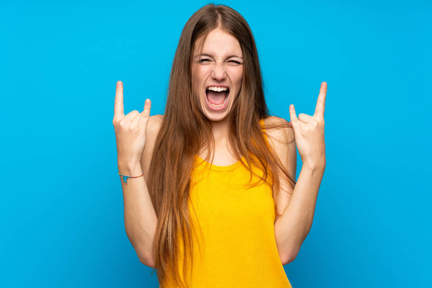 Молода жінка з довгим волоссям над ізольованою синьою стіною робить рок жест
 - Фото, зображення