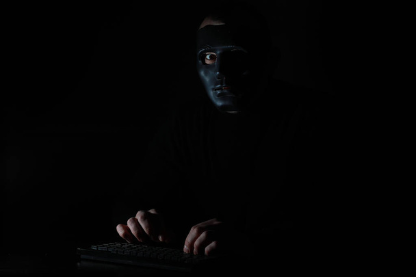 A man in a mask prints on the keyboard in the dark - Photo, Image