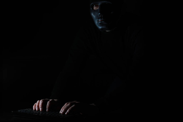 Een man in een masker drukt op het toetsenbord in het donker - Foto, afbeelding