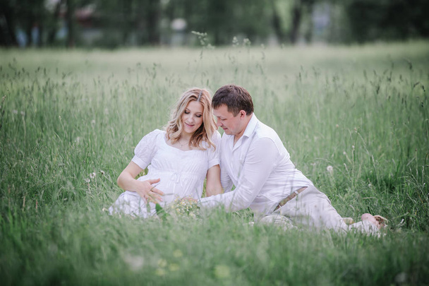 beautiful pregnant woman with her husband sitting on lawn in summer day. - Фото, изображение