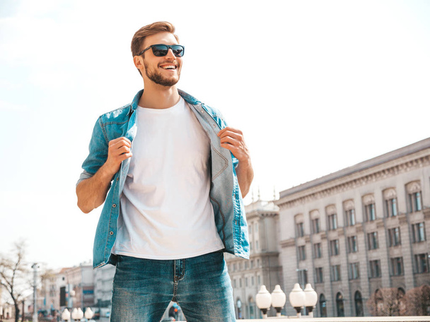 Portrait of handsome smiling stylish hipster lumbersexual businessman model. Man dressed in jeans jacket clothes. Fashion male posing on the street background in sunglasses - Foto, afbeelding