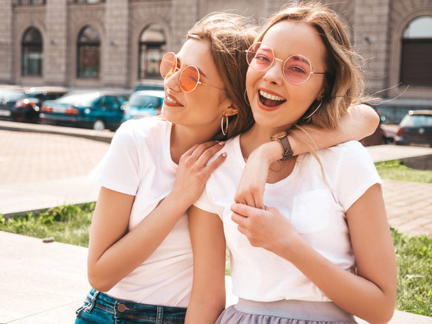 Portrait of two young beautiful blond smiling hipster girls in trendy summer white t-shirt clothes. Sexy carefree women posing on street background. Positive models having fun in sunglasses.Hugging - Foto, Imagen