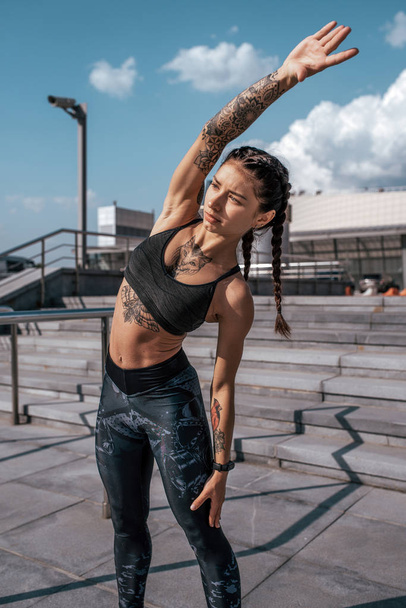 Woman athlete involved in fitness fitness workout, in summer in the city, day and morning. Active lifestyle, the concept of warming up before jogging. Sportswear leggings top. Tanned skin in tattoos. - Foto, afbeelding