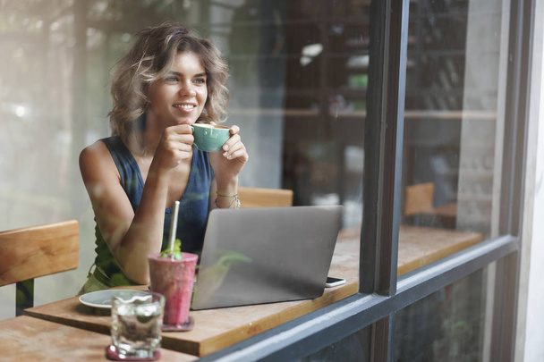 Attractive stylish female entrepreneur working outside office sit cafe look outside window passersby dreamy smiling holding cup coffee enjoy momentum working laptop prepare project - Foto, Imagen