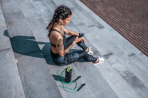 Girl athlete sitting steps resting after workout fitness summer city, day morning. Sportswear, smart phone, headphones, jumper. Active lifestyle, motivation power. Internet application, music. - Photo, Image