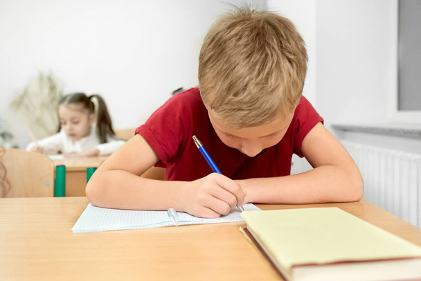 Шкільний хлопець сидить за столом, пишучи ручкою в книзі
. - Фото, зображення