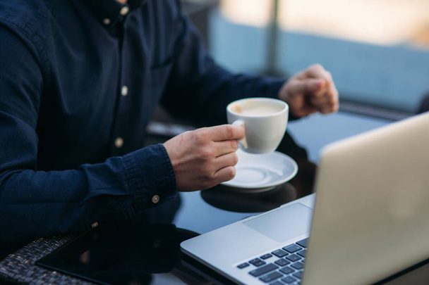 Όμορφος άνθρωπος πίνει cappuchino στο καφέ στη βεράντα και να κοιτάξει στο laptop του - Φωτογραφία, εικόνα