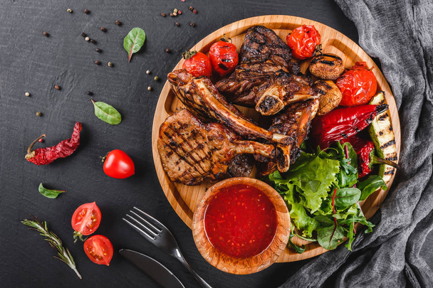 Grilled Ribeye Steak on bone and vegetables with fresh salad and bbq sauce on cutting board over black stone background. Hot Meat Dishes. Top view, flat lay - 写真・画像