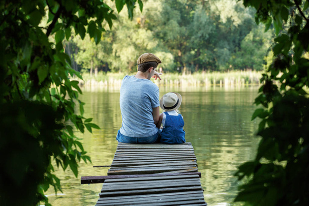 Tata i malutka dziewczynka w ogrodniczki relaks nad rzeką. Widok z tyłu,  - Zdjęcie, obraz