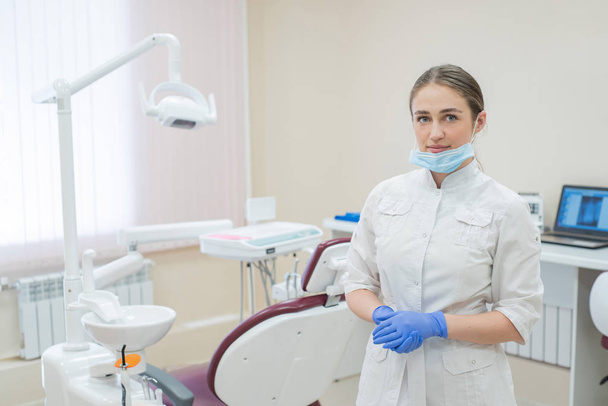 Attractive smiling female doctor in a white uniform at the workplace. A young beautiful female dentist in mask and gloves is standing in the office by the chair - Fotografie, Obrázek