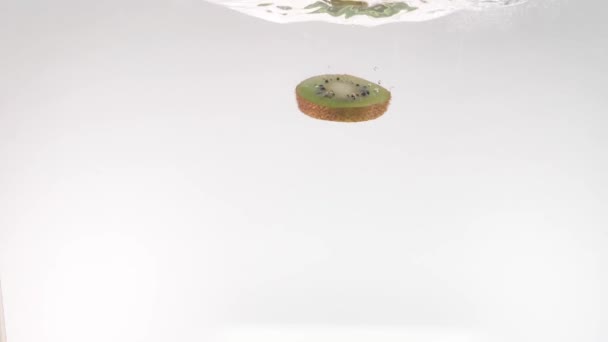 slices of kiwi fruit slowly fall into the clear water on a white background - Materiał filmowy, wideo