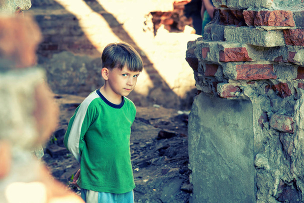 Poor and unhappy orphan boy, stands in a ruined building and looks out with danger. Staged photo. - Photo, Image