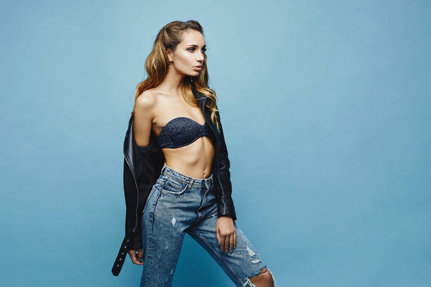 Beautiful young model woman with sexy full lips and with perfect body in a leather jacket and in fashionable jeans posing at the blue background, isolated - Φωτογραφία, εικόνα