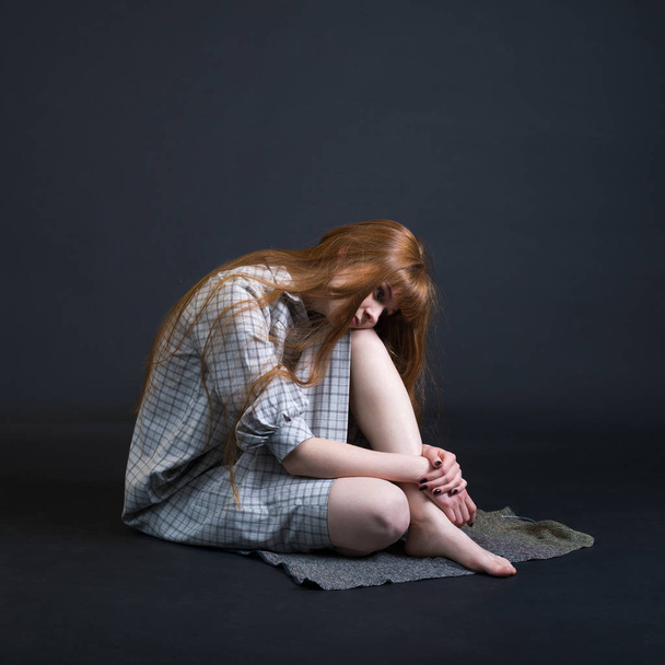 Rudowłosy młody smutna kobieta siedzi na podłodze izolowane na ciemnym tle. - Zdjęcie, obraz