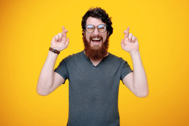 Happy bearded guy is looking at camera while crossing finger for more luck, standing over yellow background  - Photo, Image
