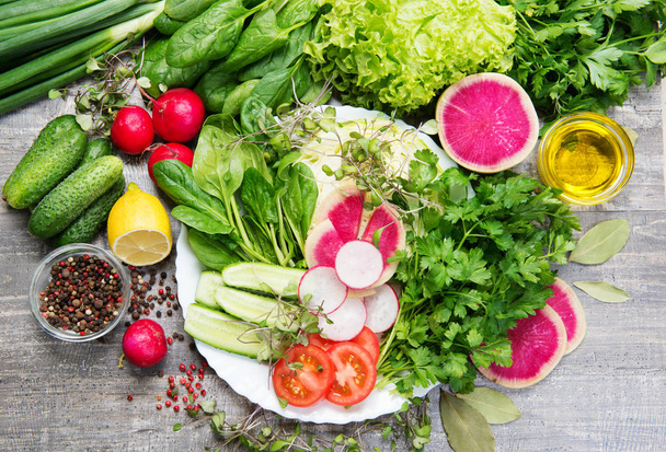 Целые витамины органической веганской пищи. На белой тарелке шпинат, редис, помидор, огурец. Рядом с другими травами и овощами
 - Фото, изображение